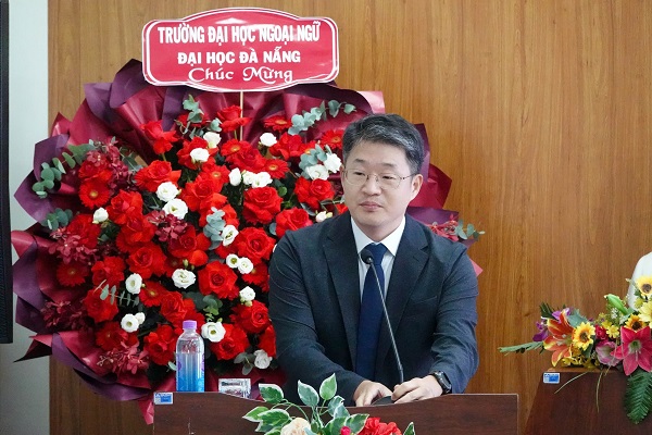 A person in a suit and tie sitting at a podium with flowersDescription automatically generated